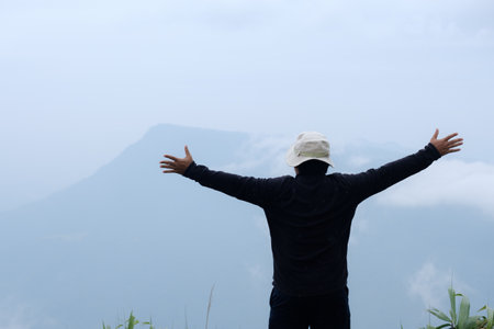 A mountaineer climber in hat stands with his arms outstretched to convey success and freedom atop a misty high mountainの写真素材
