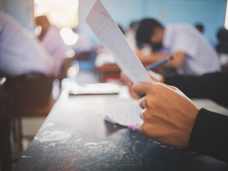 University student holding pen or pencil writing on paper answer sheet sitting on chair lecture final exam participates in exam room or classroom in uniform studentの写真素材