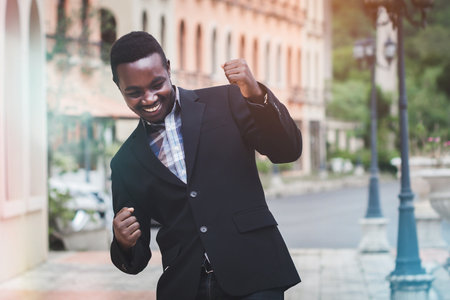 African businessman in a suit happy smile with raised hands up clenched fists on a building background. The Celebrate success and joy of winning in business conceptの写真素材