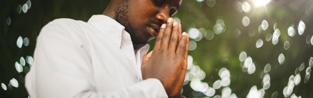 Peaceful African Man Praying In White Shirt Holding Clasped Hands In Prayer Gesture Standing With Eyes Closed Posing In Green Nature Background. Portrait Of Black Man Sending Prayers To Godの写真素材