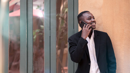Smart African American businessman in a suit stands outside near an office center and uses a telephoneの写真素材