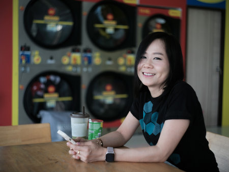 A dark-haired Asian woman sits on her cell phone with a drink next to her while waiting at a coin-operated laundry machineの写真素材