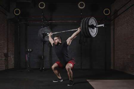 Man doing squats with heavy barbell overhead at the gym. Handsome man practicing functional training powerlifting exercisesの写真素材