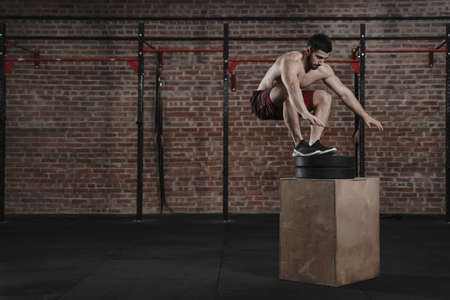 Young cross fit athlete doing box jumping at the gym. Man practicing functional training. Place for textの写真素材