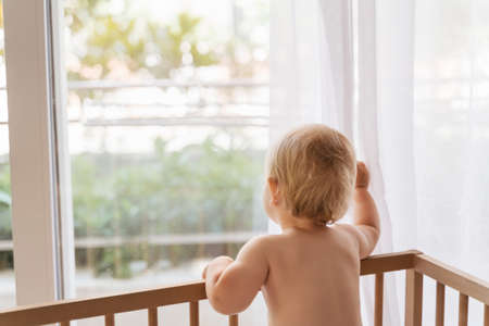 Back view of sad lonely baby standing in bed looking through window waiting for his momの写真素材