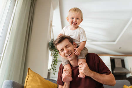 Laughing father giving his kid piggyback ride on shoulders while baby boy pulling daddys hair