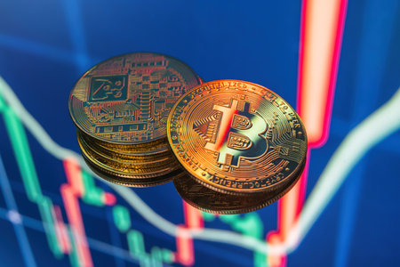 Closeup of stacked golden bitcoins lying on blue digital display with crypto candlestick chartの写真素材