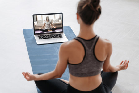 Close up of young sporty woman practicing yoga online with laptop at home. Yoga instructor conducting video training via video conference. Meditation, Sukhasana, relax. Healthy lifestyle conceptの写真素材