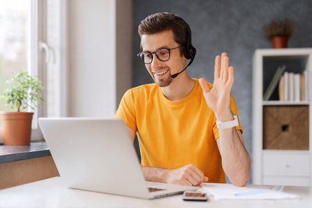 Guy wearing headset, start lesson, looking at laptop screen, waving hand, greeting tutor, has online class via video call, get skills through internet, remote education distantly with modern techの写真素材