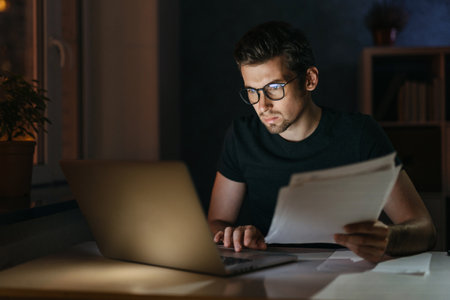 Portrait of tired focused young man in glasses working remotely late at night at home office with computer, looking at laptop, thinking about deadline. Overwork, negative thoughts, deadline conceptの写真素材