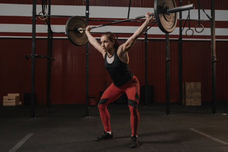 Young crossfit female athlete lifting heavy barbell overhead at the gym. Strong woman doing functional training. Lifting weights.の写真素材