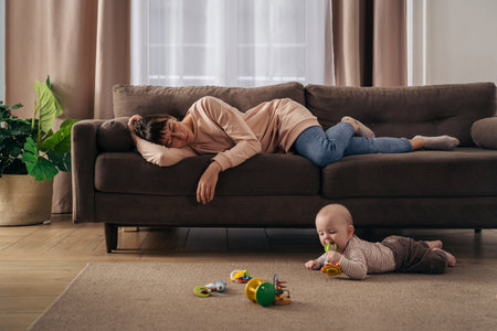 Young tired mother suffering from lack of sleep, sleeping on sofa while her little infant baby is playing on floor. Exhausted mom experiencing postnatal depression, does not want to play with her sonの写真素材