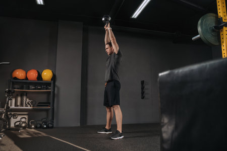 Handsome athletic young man lifting kettlebell with two hands as crossfit workout. Muscular man holding kettlebell overhead at gym.の写真素材