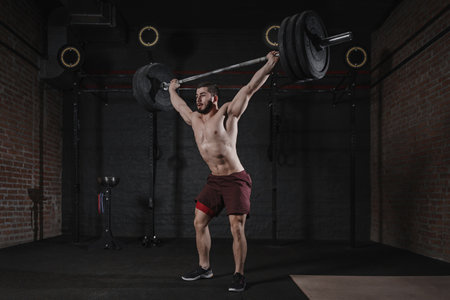 Crossfit athlete lifting barbell overhead at the gym. Shirtless man doing functional training....