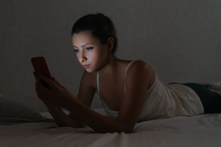 Young woman messaging with smartphone in bed at night. Social network dependence. Gadget addict concept. Harm of using phone before going to bedの写真素材