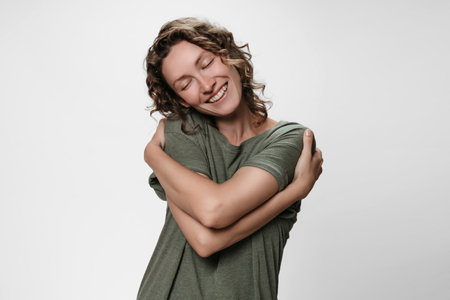 Young curly woman hugging herself, looks happy, loves herself, has high self esteem, smiles from pleasure. Isolated over white. Self love and self care concept.の写真素材