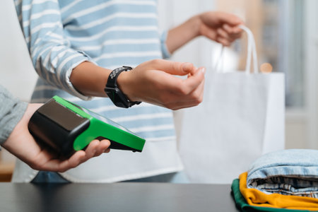 Modern woman using terminal for contactless payment with smartwatch on counter in clothing store. NFC technology. Female customer paying for purchase through smart watchの写真素材