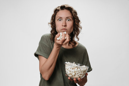 Young woman with curly hair eating popcorn, watching a movie or TV shows with great surprise, stare at TV, isolated on white background. Copy spaceの写真素材