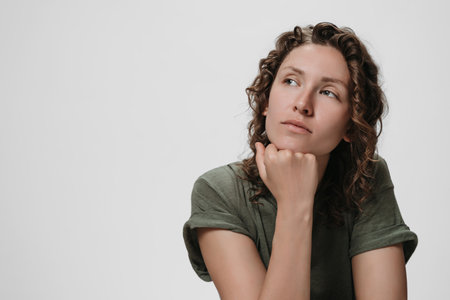 Young curly caucasian woman keeps hand under chin, being deep in thoughts, thinks about something....