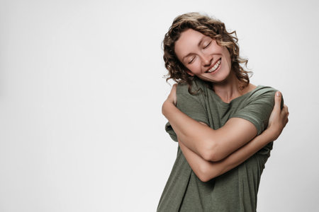 Young curly woman hugging herself, looks happy, loves herself, has high self esteem, smiles from pleasure. Isolated over white, free copy space for your text. Self love and self care concept.の写真素材