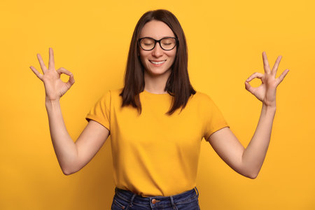 Mindful peaceful young woman with closed eyes meditates indoor on yellow wall, keeps hands in mudra gesture, relaxing after long hours of hard working, holds fingers in yoga sign. Relaxing conceptの写真素材