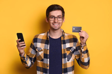 Young caucasian man in glasses using mobile phone and credit card for online shopping, e-commerce, online banking, smiling isolated over yellow studio background. Technology and reaction conceptの写真素材
