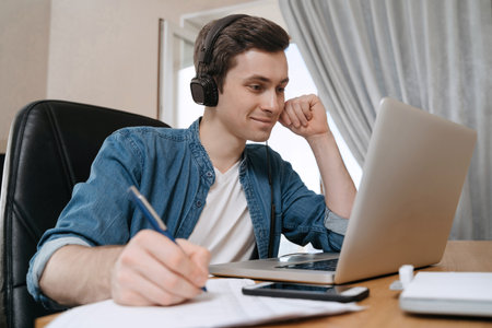 Happy guy student wearing headphones using laptop for distance education, enjoying conference video call, watching webinar, listening audio course, writing notes, studying with teacher, having lessonの写真素材