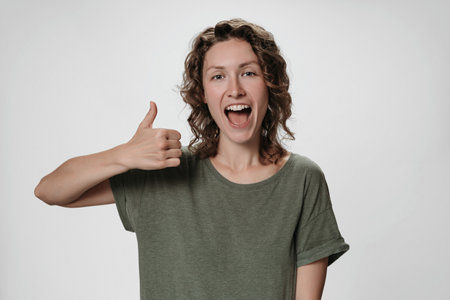 Cheerful enthusiastic young curly woman with opens eyes and mouth widely showing thumbs up gesture to camera, expression her like and approval of idea or project, smiling broadlyの写真素材