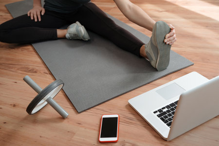 Young female in sport wear watching online yoga or workout lesson on laptop and doing exercises on floor while staying at home during coronavirus pandemic. Fitness and wellness conceptの写真素材