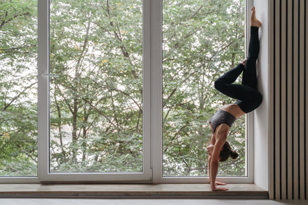 Attractive yoga girl standing on hands against wall in daylight near big window. Fit woman...