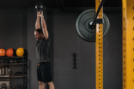 Muscular weightlifter doing an exercise with kettlebell at dark crossfit gym with professional sport...