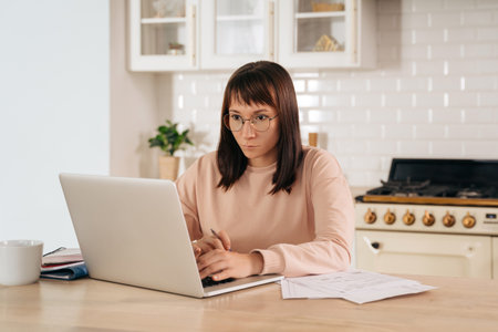 Serious woman wearing glasses working on laptop online, sitting at table in kitchen, looking at computer screen. Focused female using internet for distance education, searching information, up-skillの写真素材
