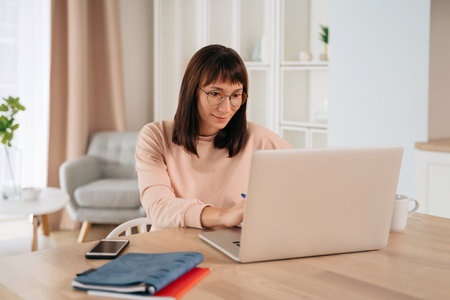 Smiling young woman wearing glasses working remotely, typing on laptop, looking at computer screen, sitting at desk in her home office. Female enjoying elearning process, studying online at homeの写真素材