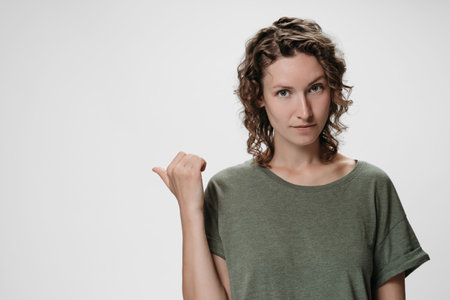 Caucasian curly woman with friendly expression, points aside with thumb, shows free copy space for your advertising content on white background. Advertisement conceptの写真素材