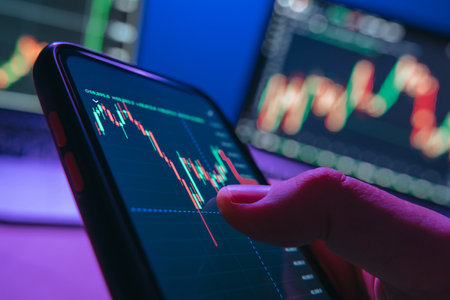 Close up of trader hand holding smartphone, using trading application analyzing crypto and stock market trends at his neon lit office with multiple monitors on backgroundの写真素材