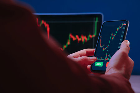 Trader hands holding smartphone, using trading app for analyzing financial, stock market and cryptocurrency data, with red and green candlestick charts on screen.の写真素材