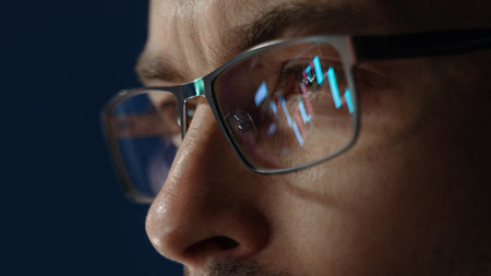 Close up of focused crypto trader wearing glasses with stock market indexes reflecting in lenses while he analyzing cryptocurrency market in dark office. Trading and investment in cryptoの写真素材