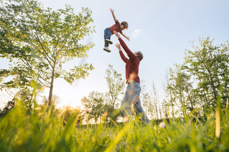 Joyful father tossing young son into the air, playing in a sunny park, surrounded by trees and fresh green grass at sunset. Fathers day and family bonding concept. Copy spaceの写真素材