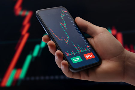 Close up, trader holding smartphone with a mobile trading platform application on screen, with candlestick chart and financial graphs for stock or crypto investments on background.の写真素材