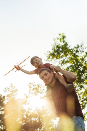 A father carries his excited son on his shoulders in a park, the boy holding a toy airplane and imagines flying as they enjoy a fun, carefree moment together. Copy spaceの写真素材