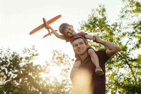 Smiling father carries little son on shoulders as boy holds toy airplane, imagining he is flying, enjoying playful time together in a sunny park. Fatherhood and fathers day concept. Space for textの写真素材