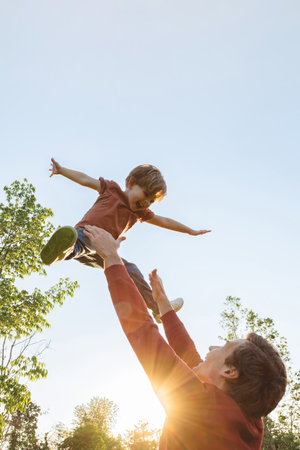 Father tossing his laughing joyful son into the air against a bright sky in a park on a sunny day. Cheerful boy playing with his dad outdoors, creating happy childhood memories together. Copy spaceの写真素材