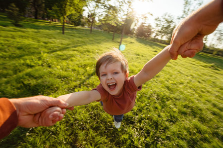 First-person view, father holding his sons hands, spinning him around in a circle at the park, joyful child laughs, enjoying the playful moment, creating happy childhood and family memories togetherの写真素材