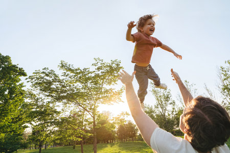 A cheerful boy is tossed into the air by his dad during a fun bonding activity at the park, surrounded by warm evening sunlight. Copy spaceの写真素材