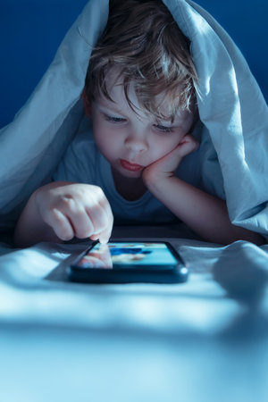 Little boy lying in bed under a blanket at night, intently watching and scrolling social media content on smartphone screen, face lit by blue light. Gadget addiction and overuse. Copy spaceの写真素材