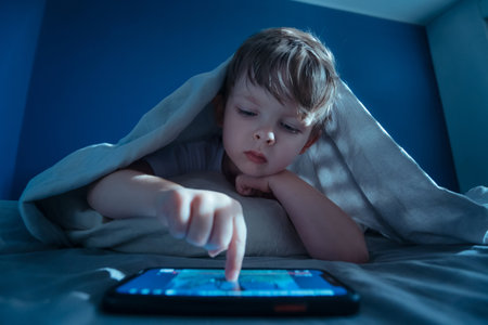 A little boy lies under the blanket late in the evening, uses smartphone, pressing play on screen to watch next cartoon. Digital addiction and prolonged gadget overuse.の写真素材