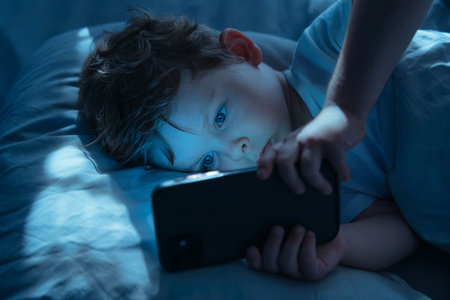Little boy lies in bed at night, staring intently at his smartphone screen. Child watches cartoons on his gadget, illuminated by blue light of the screen. Childhood and technology, Digital addictionの写真素材