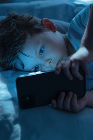 Little boy lies in bed at night, staring intently at his smartphone screen. Child watches cartoons on his gadget, illuminated by blue light of the screen. Childhood and technology, Digital addictionの写真素材