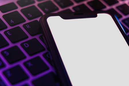 Mock up, smartphone with white blank screen lying on laptop keyboard in violet light, trendy digital concept for modern gadget, technology, or application design projects. Space for textの写真素材