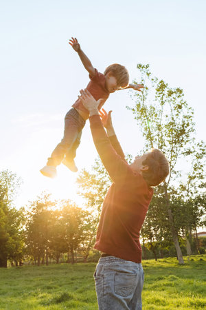 Father tossing his smiling young son into the air and catching him while playing together outdoors in a sunny park at sunset. Space for textの写真素材